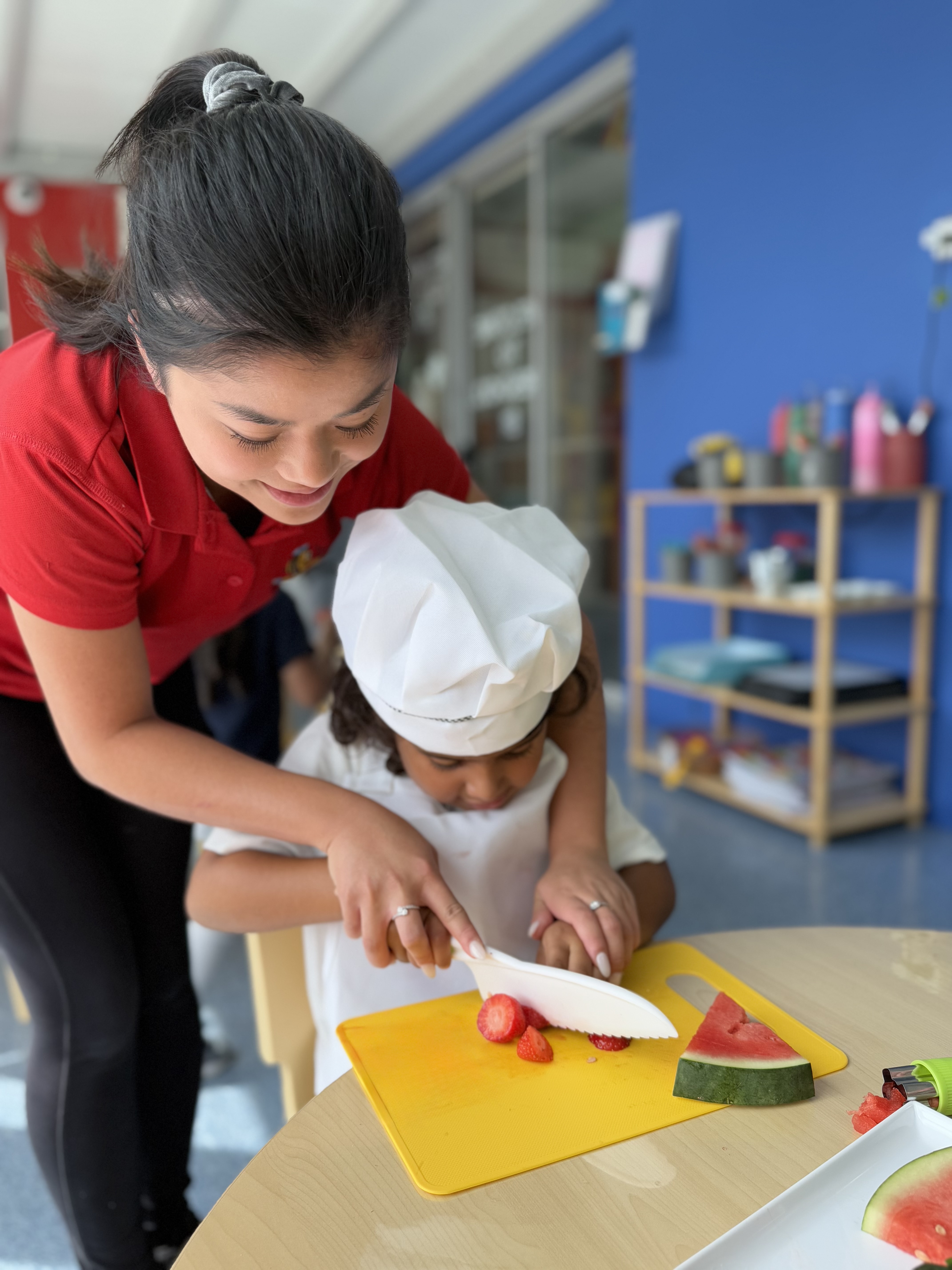 Teacher assisting a child with cutting tomatoes in a classroom kitchen setting.